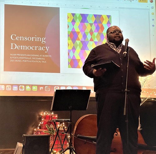 Man standing on stage in front of a microphone. Behind him on a screen are the words "Censoring Democracy: An Evening and Busboys and Poets, December 8, 2021"