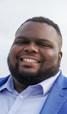 Image of a man smiling. He is wearing a suit jacket and collared shirt, and he has a beard.