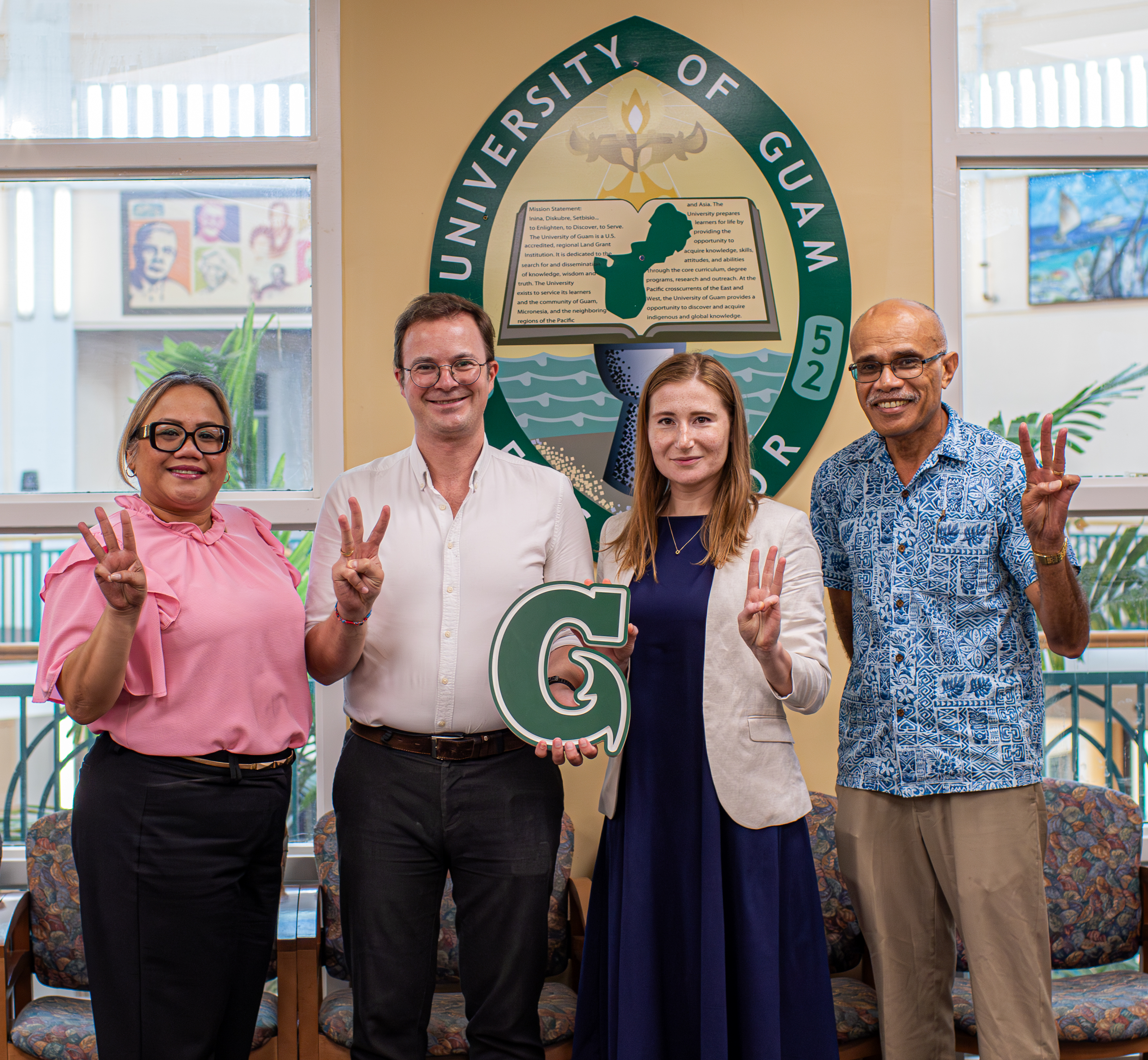 Four individuals standing shoulder-to-shoulder front of a sign that reads "University of Guam". They are all holding up 3 fingers and the 2 inner people are holding the logo of the university.