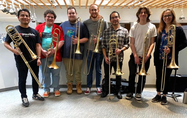 7 people standing shoulder to shoulder, smiling and holding their trombones.