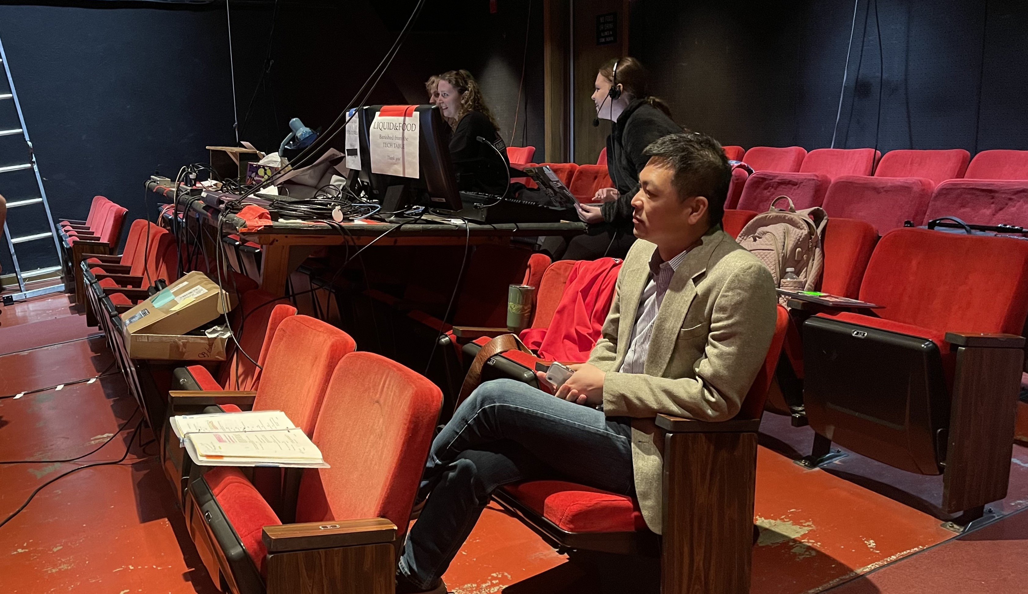 A man sitting in the aisle seat of a theatre with red chairs, looking thoughtfully towards the stage