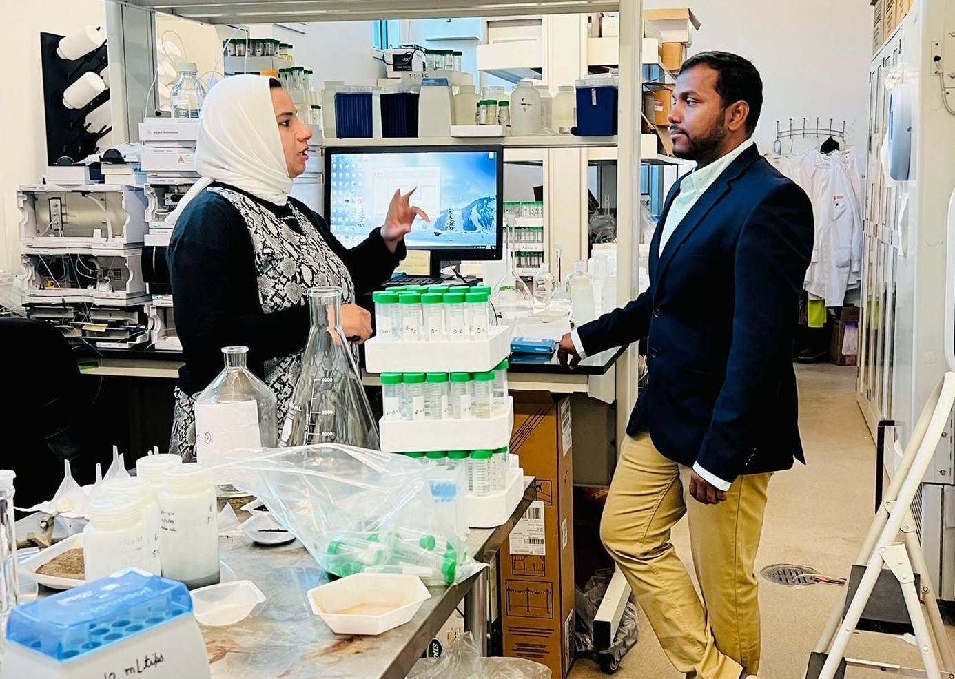 Two individuals talking in a lab, standing in front of a computer and surrounded by various stacks and cluttering of lab supplies.