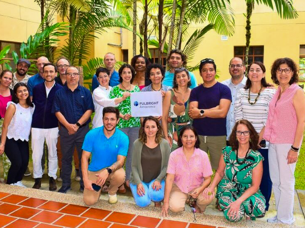 A group of twenty people poses with a "Fulbright Amazonia" banner in front of a yellow building and greenery.