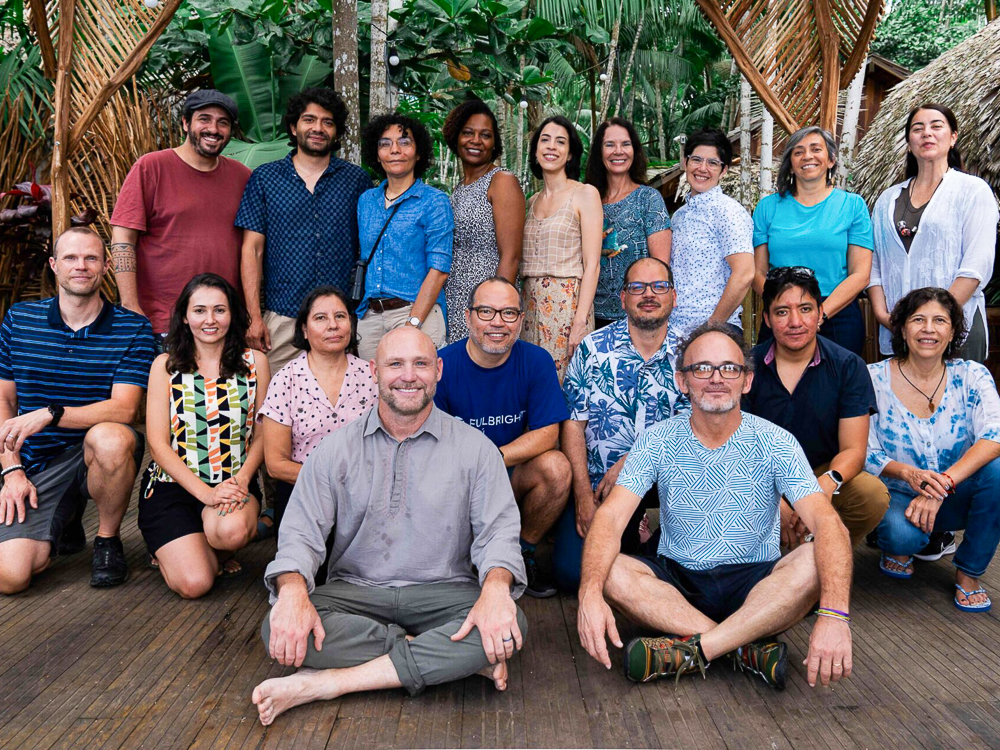 A group of sixteen people posing together outdoors in a tropical setting with greenery and wicker elements.