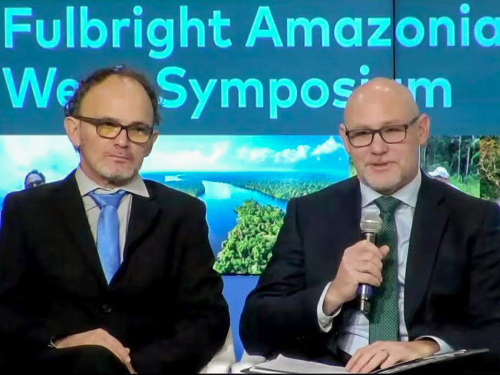 Two men seated at the Fulbright Amazonia Week Symposium, with an Amazonian landscape backdrop.