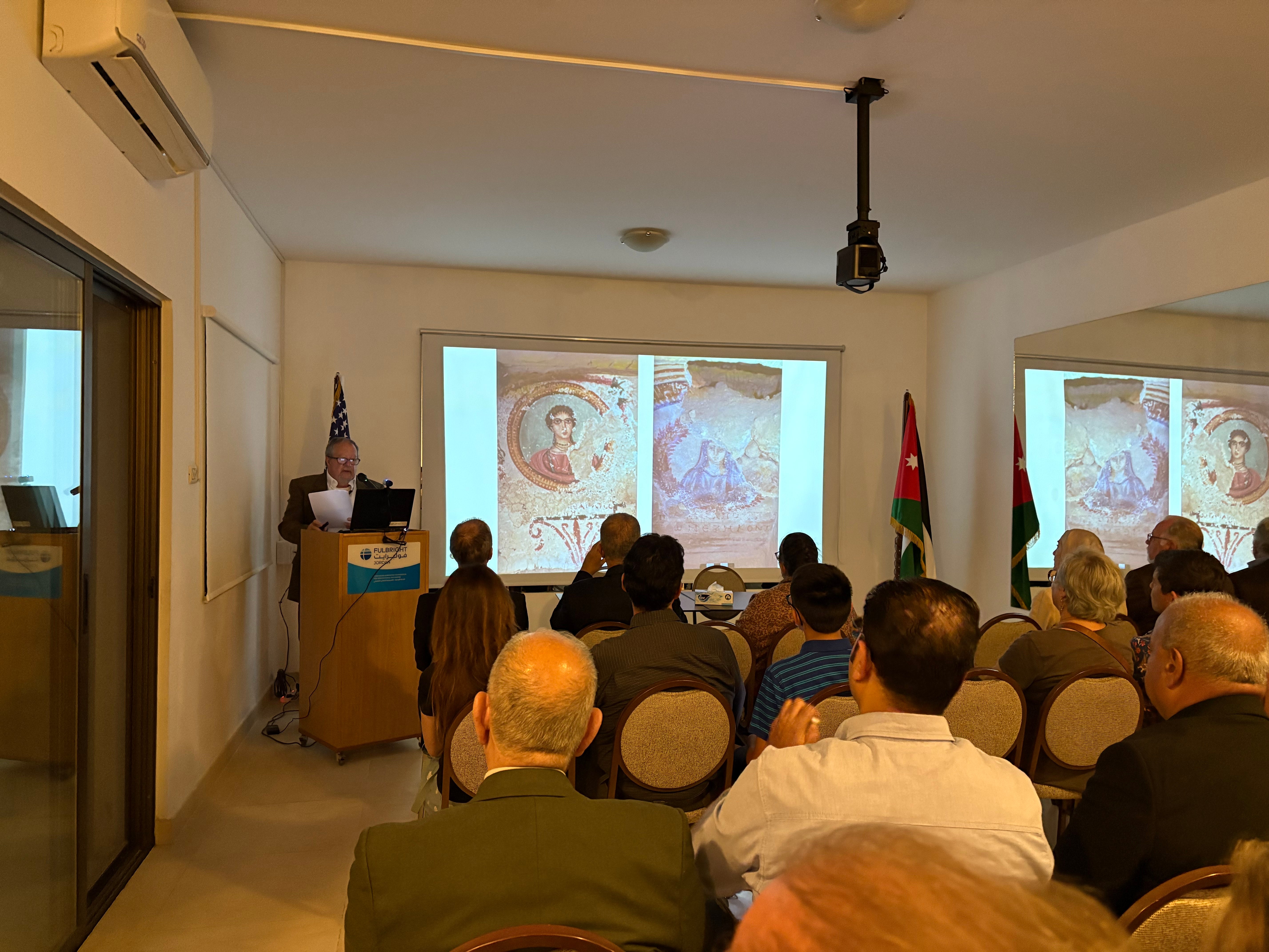 A speaker presenting in front of a projected image of mosaics in a conference room with an attentive audience.