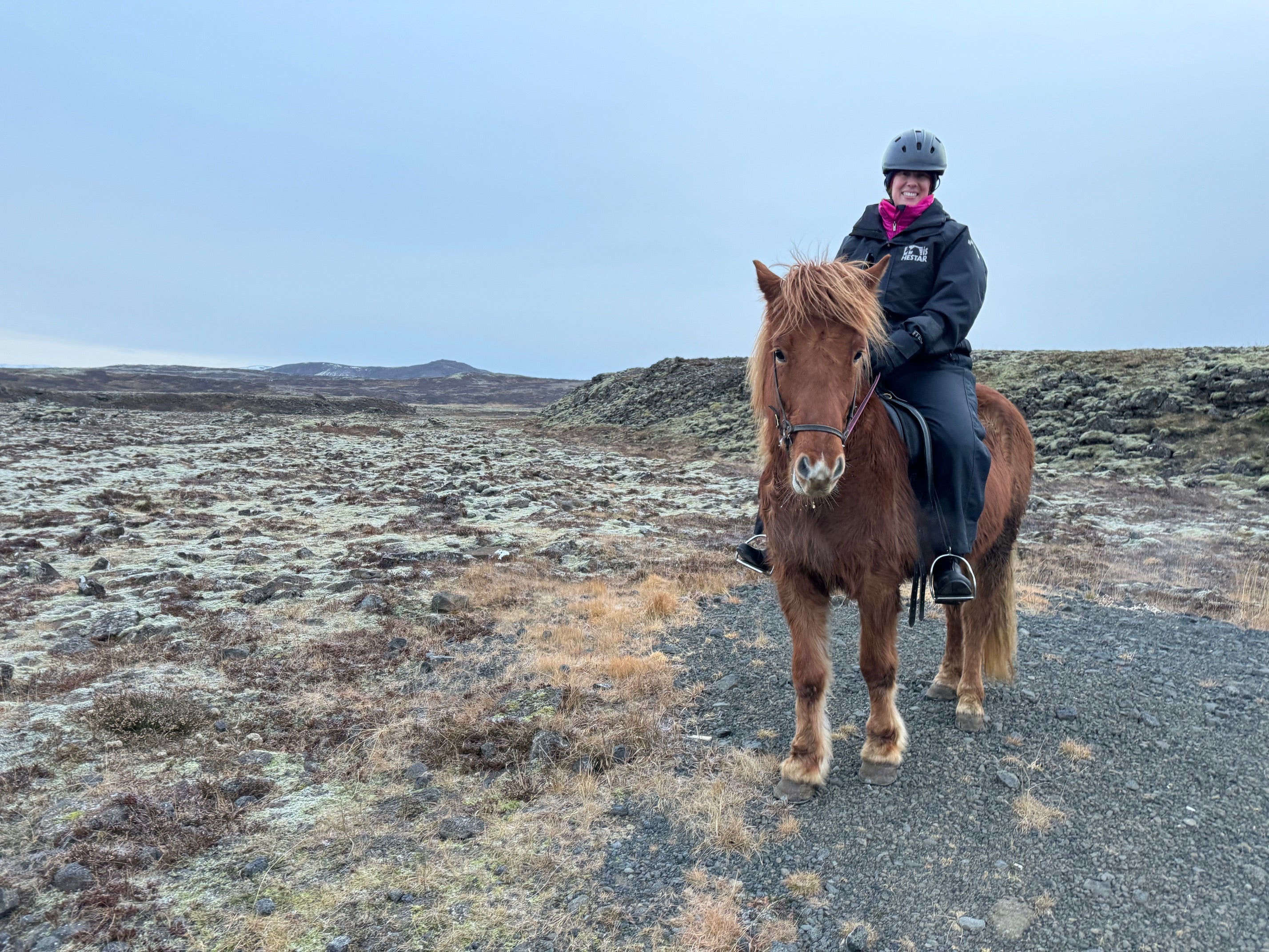 Person in a black outfit and helmet rides a brown horse on a rocky, moss-covered landscape. The scene is calm with an overcast sky.