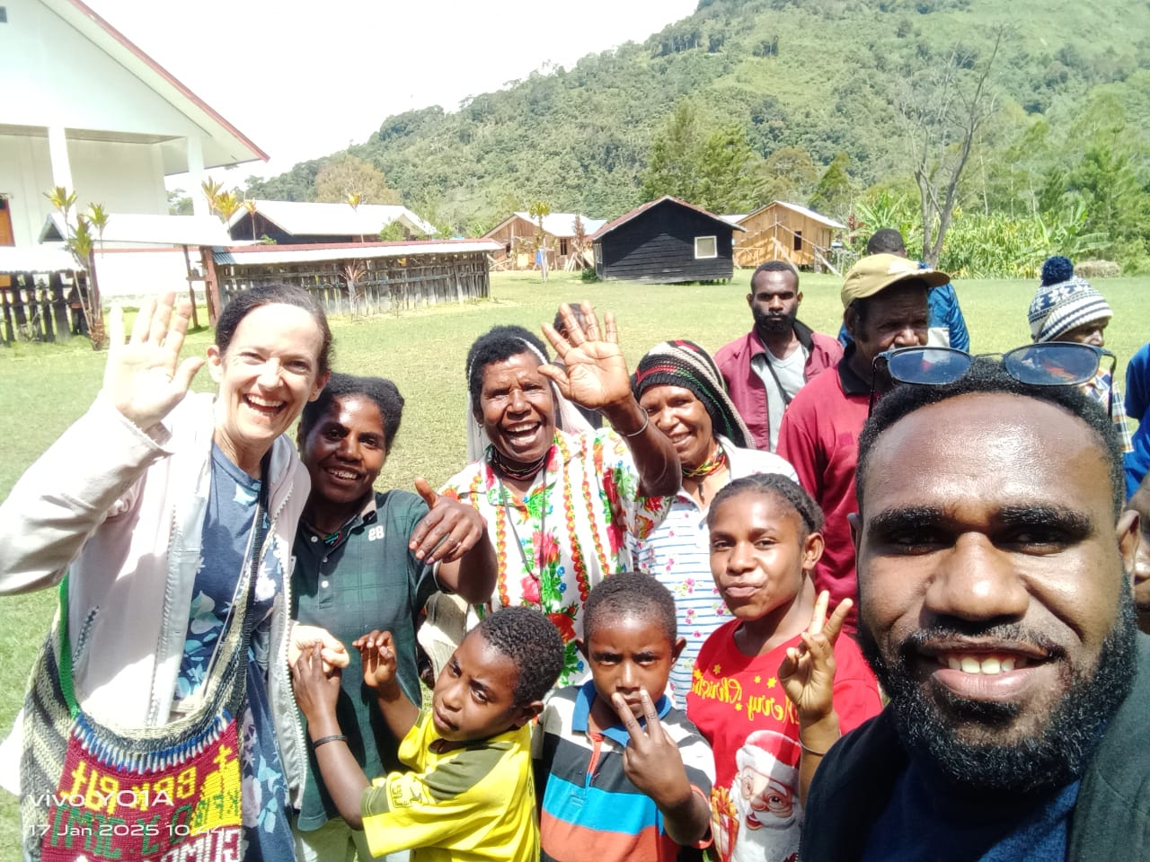 A group of people smiling and waving outdoors with hills and wooden buildings in the background.