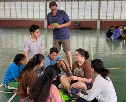 A man stands holding colorful cards, speaking to a seated group in a gym.