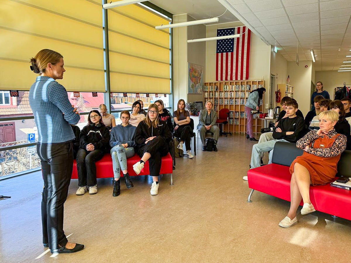 A woman speaks to a seated group in a classroom with large windows and an American flag displayed.