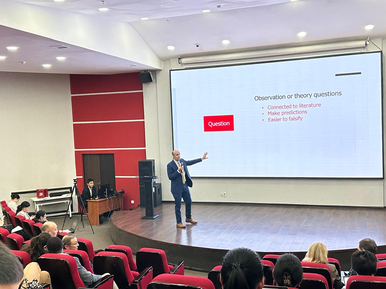 A lecturer presenting in a modern lecture hall with a large screen displaying a slide on "Observation or theory questions."