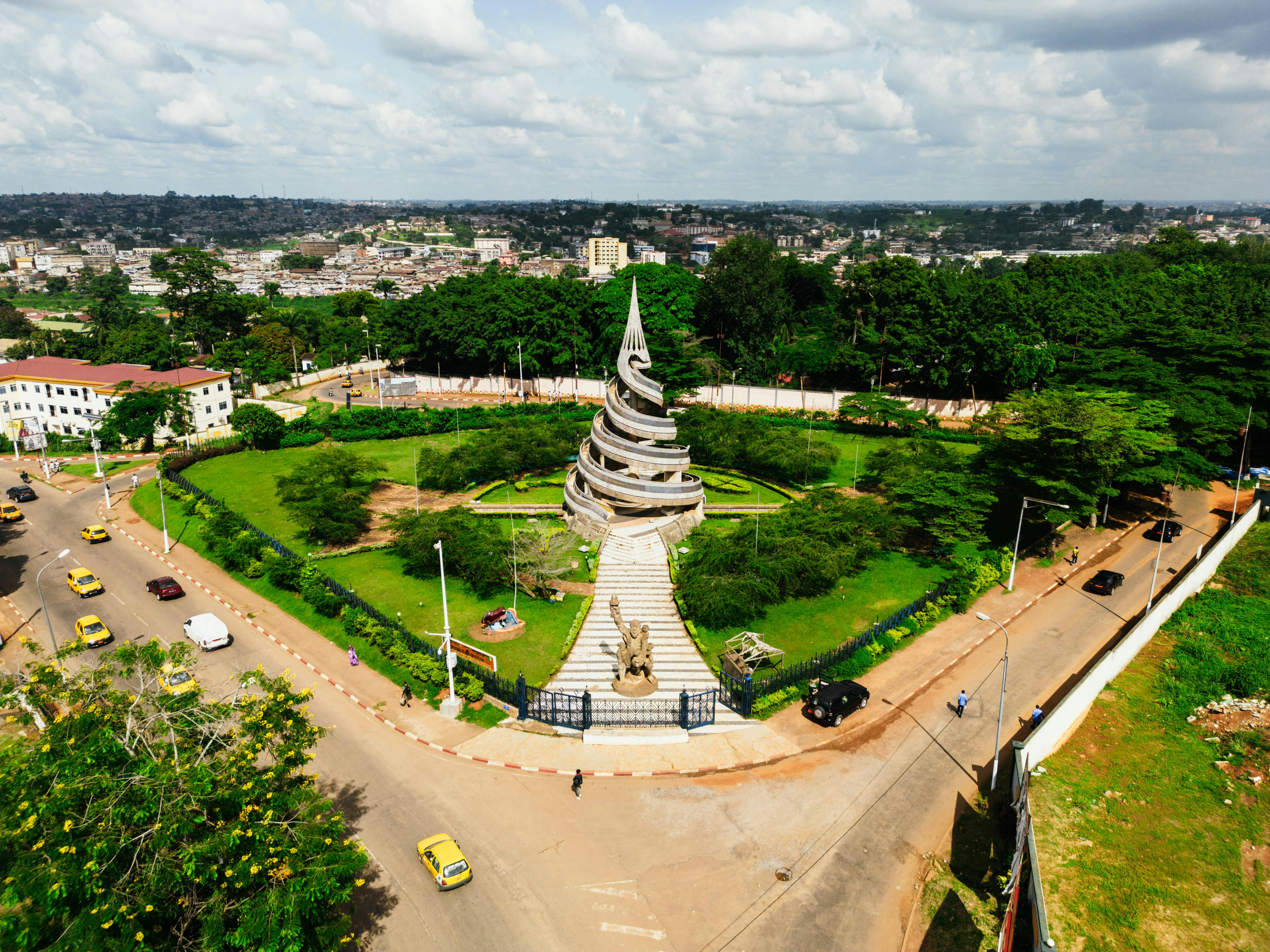 Aerial view of a spiraling monument in the center of a green, circular park. Surrounded by roads with yellow taxis, cityscape in the background. Bright, sunny day.