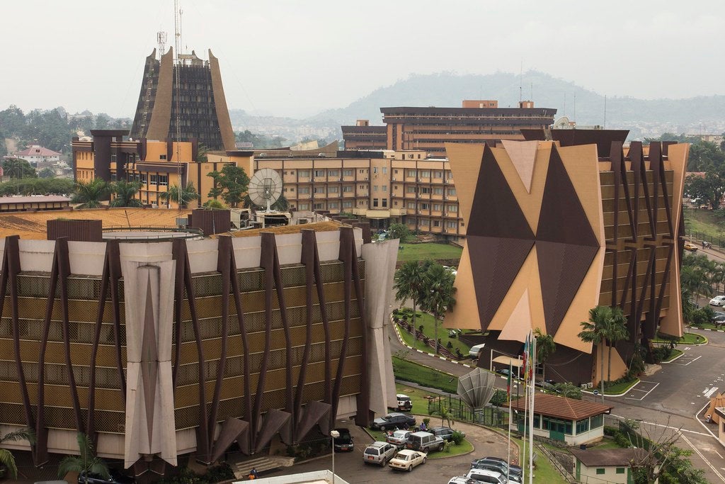 Aerial view of modern government buildings with geometric designs in warm tones. Lush greenery and parked cars surround the structures, exuding a formal yet vibrant atmosphere. In the background, a hilly landscape fades into the misty sky.