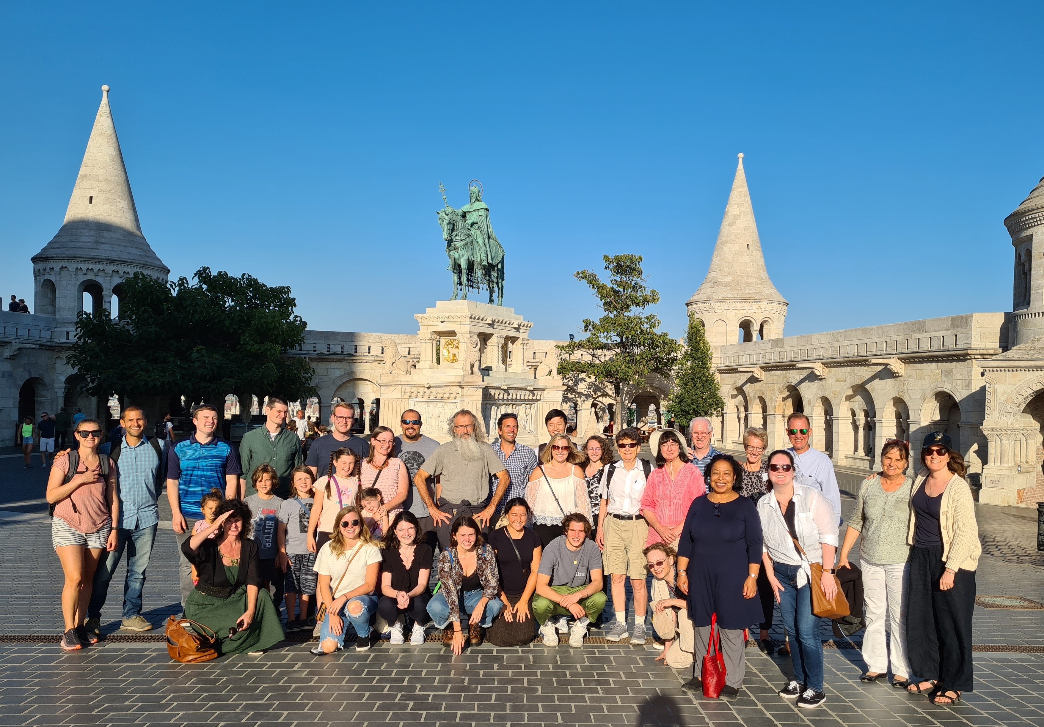 A group of people pose in front of a historical monument in Budapest, featuring a mounted statue and conical towers, under a clear blue sky.