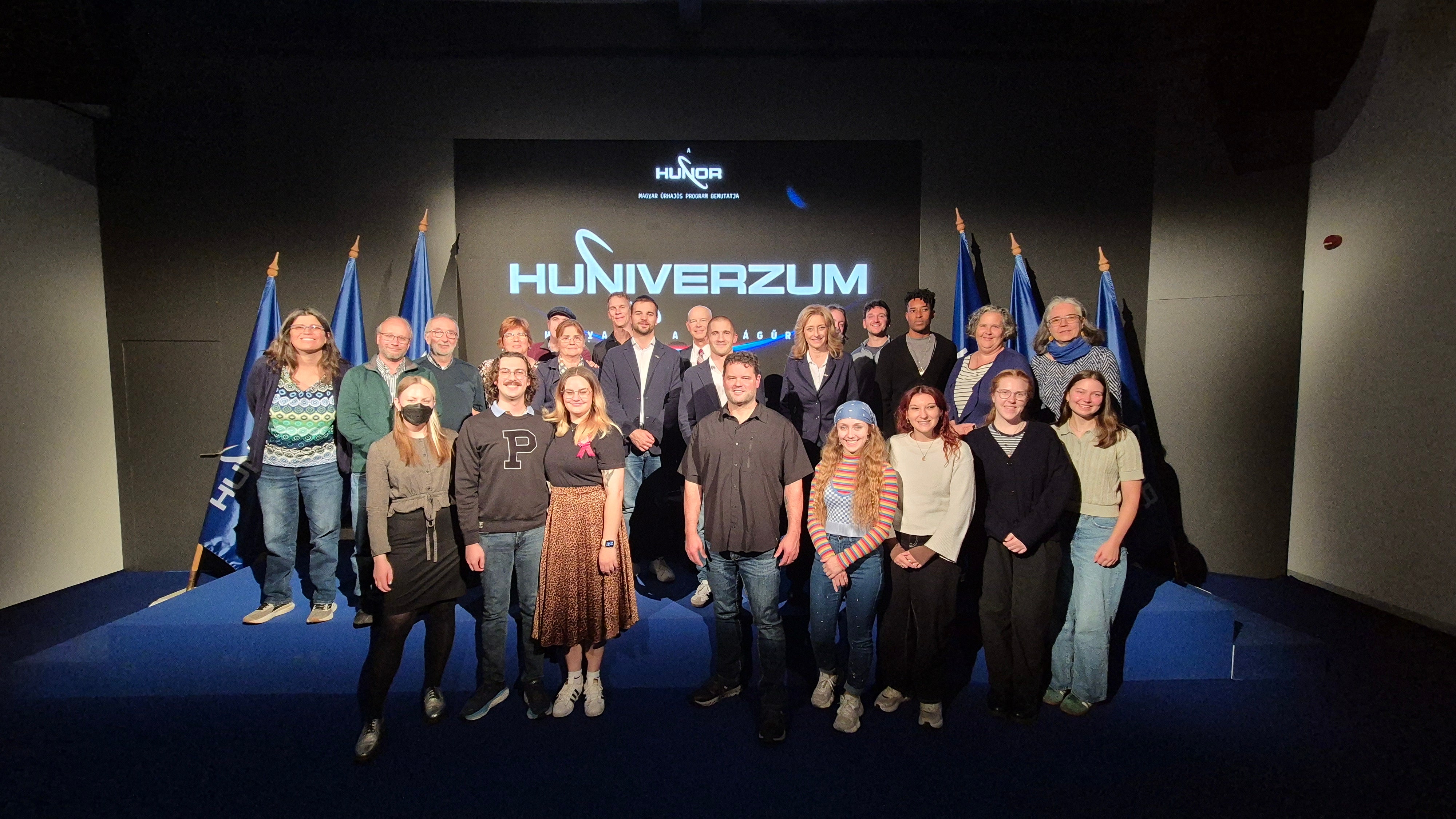 A group of about twenty people posing in front of a backdrop with the word "HUNIVERZUM."