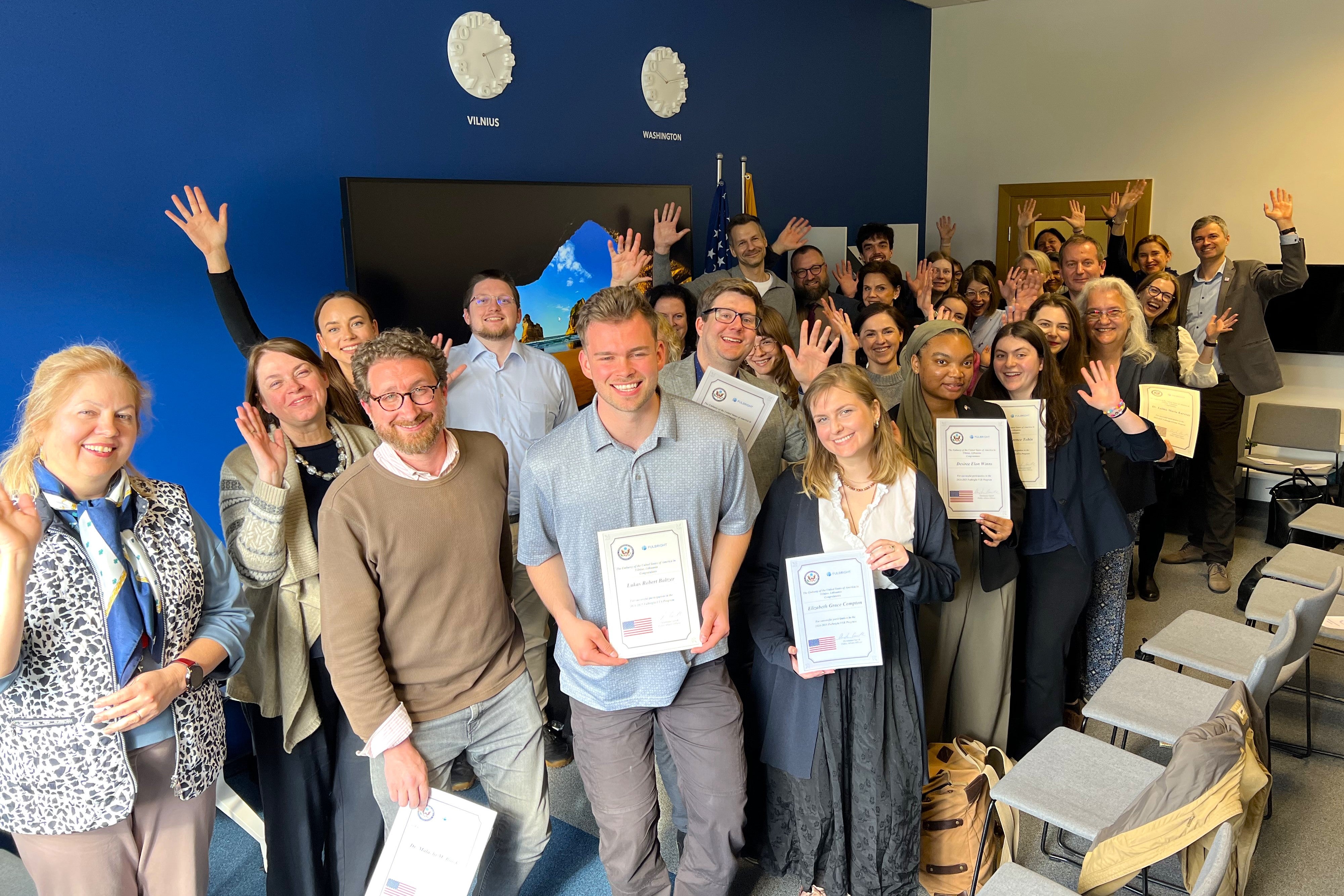A group of smiling people holding certificates in a room with blue walls and two clocks labeled "VILNIUS" and "WASHINGTON."
